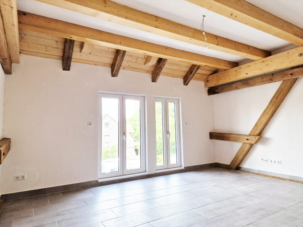 Heller Wohnraum im Dachgeschoss mit sichtbaren Holzbalken, großen Fenstern und moderner Innendämmung hinter verputzten Wänden