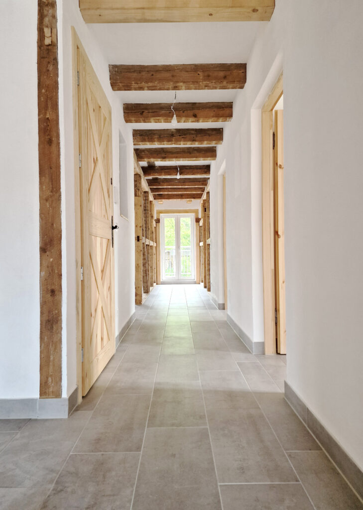 Flur im sanierten Gebäude mit sichtbaren historischen Holzbalken an der Decke, hellem Fliesenboden und Holztüren im Landhausstil